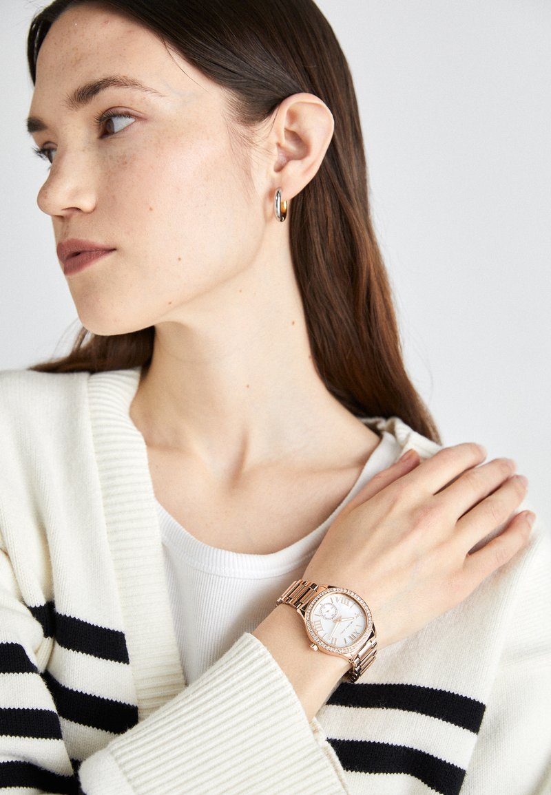 Reloj de oro rosado con un dial blanco redondo, números romanos y una pulsera de metal. Lleva puesto en una muñeca, acompañado de un cárdigan texturizado en color crema.