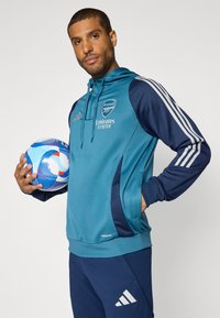 Teal and navy athletic hoodie with logo and text "Emirates Fly Better." Paired with navy pants featuring white Adidas logo and holding a multicolored soccer ball.