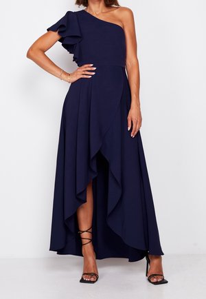 Kobieta w granatowej, asymetrycznej sukience na jedno ramię z falbanką, z asymetrycznym dołem, nosząca czarne sandały na wysokim obcasie z paskami, stoi z ręką na biodrze.