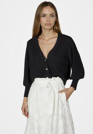 Frau mit langen braunen Haaren, die eine schwarze, mit Knöpfen versehene Strickjacke mit Ballonärmeln und einen hoch taillierten weißen Rock mit Stoffgürtel trägt.