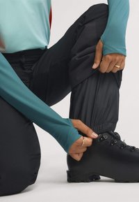 Čierne turistické topánky s kovovými očkami, nosené s čiernymi nohavicami a tyrkysovou košeľou s dlhým rukávom, prispôsobujúce elastický lem nohavíc.