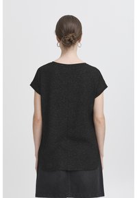 Seljapidi nähtav pruunide juustega naine, kellel on kuklas soeng, kandmas rõngakujulisi kõrvarõngaid, musta täpilise mustriga lühikeste varrukatega pluusi ja tumedat seelikut.