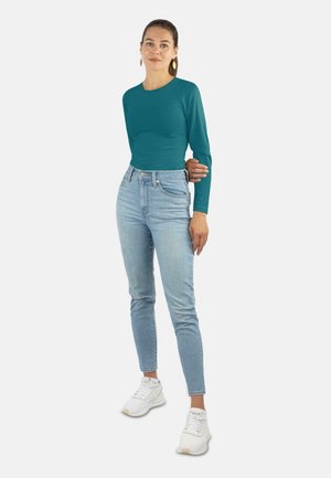 Frau steht mit Blick nach vorne, trägt ein türkisfarbenes Langarmshirt, hellblaue Jeans und weiße Turnschuhe, mit nach hinten gebundenem Haar vor einem schlichten Hintergrund.