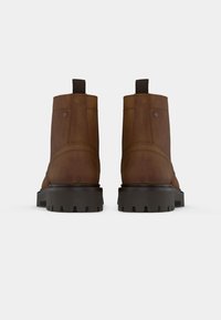 Botas de piel marrón hasta el tobillo con una textura suave, tirador y suela negra resistente con tacos pronunciados; diseñadas sin hardware visible.