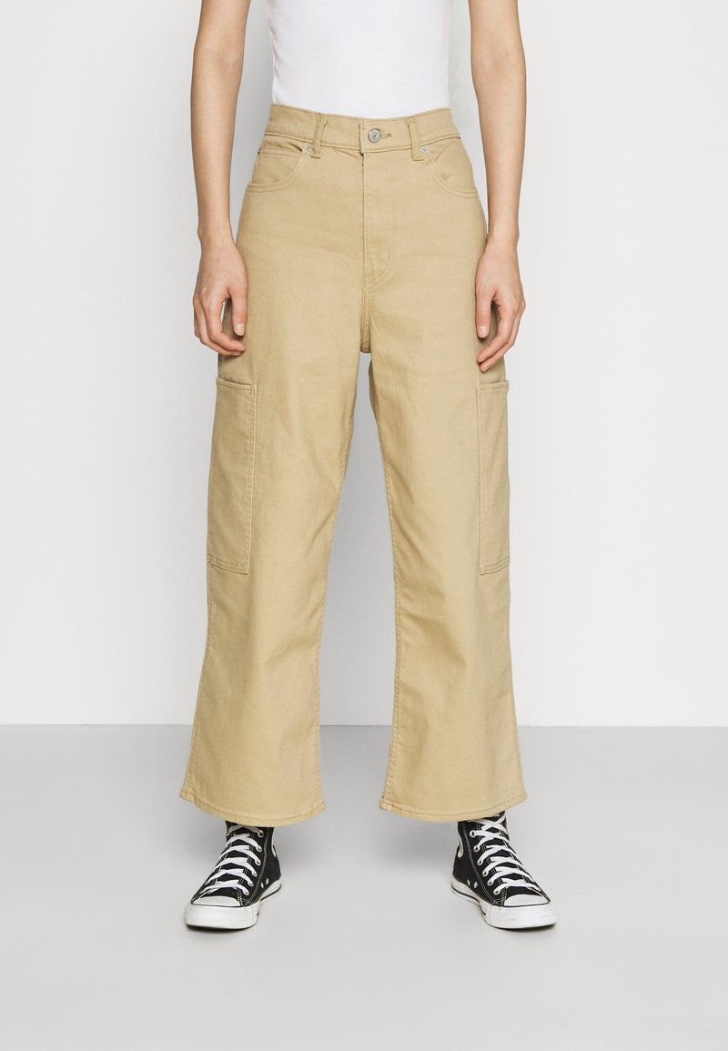 Pantalones cargo beige de pierna ancha con bolsillos delanteros, confeccionados en tela de algodón, con cintura alta y diseño de pierna recta. Combinados con zapatillas negras.