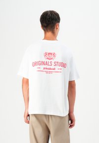 White cotton t-shirt with short sleeves, featuring a red graphic print on the back that includes the text "ORIGINALS STUDIO" and a crest design.