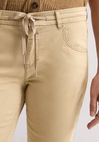 Close-up van beige broeken met voorkeurszakken en een strikband van stof rond de taille en een hand.