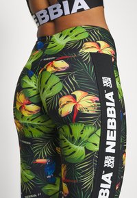 Legginsy fitnessowe z kolorowym tropikalnym wzorem, obejmującym zielone liście i kwiaty, czarne akcenty po bokach oraz białe logo. Wykonane z elastycznego materiału.