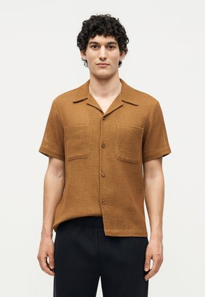 Νέος άνδρας με σγουρά σκούρα μαλλιά, φοράει κοντομάνικο καφέ πουκάμισο με υφή και μαύρο παντελόνι, στέκεται μπροστά σε λευκό φόντο.