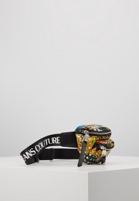 Borsa in tessuto multicolore con borchie, chiusura con zip e tracolla nera con l'etichetta "JEANS COUTURE." Forma compatta e rotonda con motivi floreali.