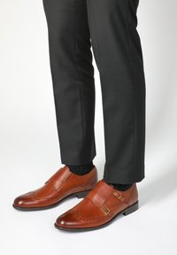 Braune Lederschuhe mit strukturiertem Finish, ausgestattet mit zwei Schnallen und dekorativen Perforationen, kombiniert mit dunklen Hosen und schwarzen Socken.