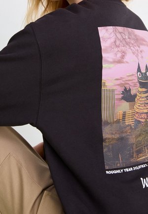 Person iført en sort sweatshirt med en farverig grafik af et tegneseriemonster i et bylandskab ved solnedgang på ryggen.