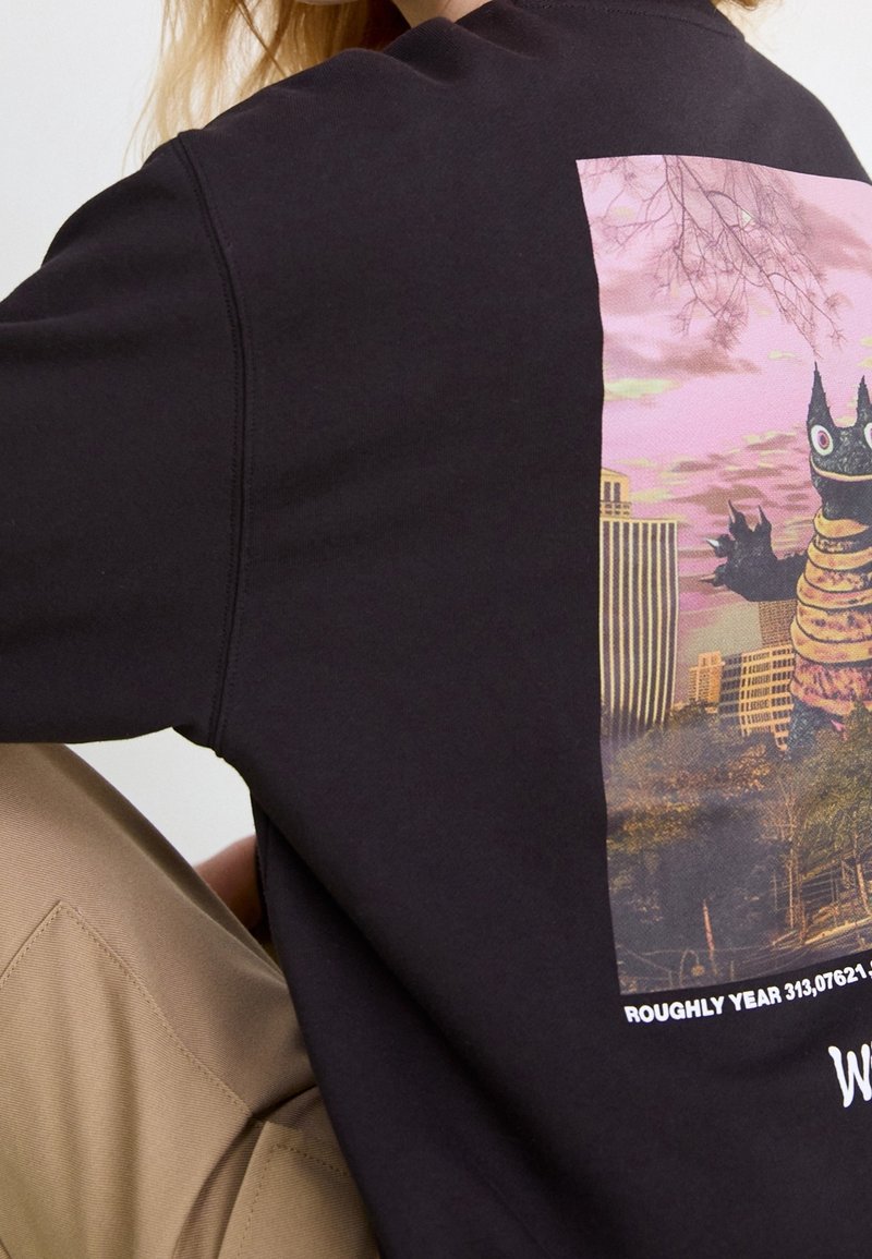 Personne portant un sweat-shirt noir avec un graphisme coloré d'un monstre de dessin animé dans un paysage urbain au coucher du soleil dans le dos.