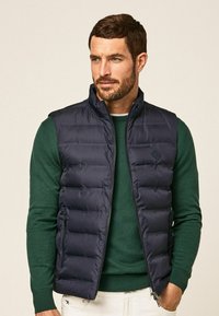 Marineblaue Steppweste mit hohem Kragen, horizontalen Nähten und seitlichen Reißverschlusstaschen, getragen über einem dunkelgrünen Pullover. Glatte Textur, leicht.