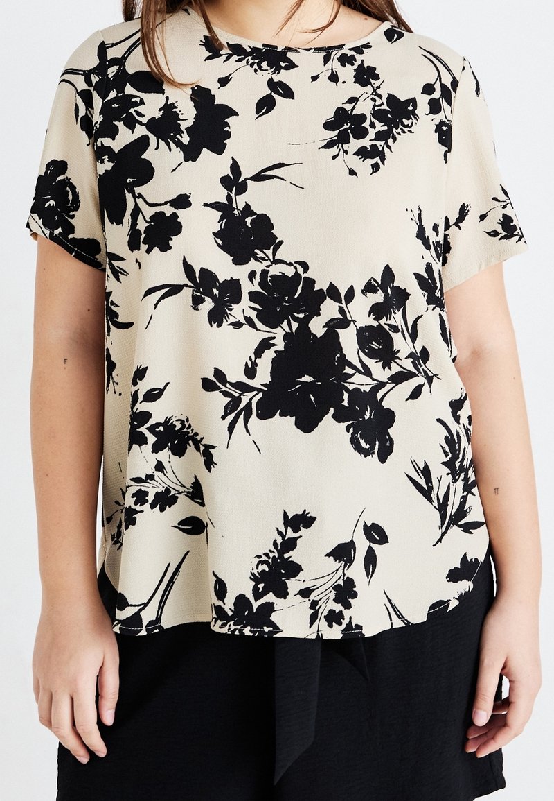 Femme portant un haut beige à manches courtes avec un imprimé floral noir et un bas noir, montrée des épaules aux hanches.