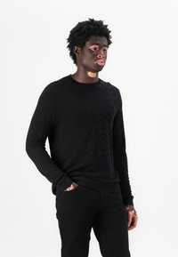 Sort strikket sweater med et struktureret mønster, rund halsudskæring og ribbet kant, parret med sorte bukser. Glat stof og tætsiddende design.