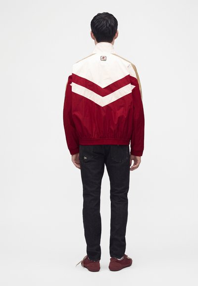 Homme portant une veste rouge et blanche bicolore, un jean noir et des baskets rouges, debout de dos devant un fond uni.