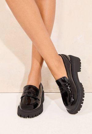 Krydsede ben iført skinnende sorte chunky loafers med tykke rillede såler mod en lys beige baggrund.