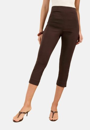 Kobieta ubrana w ciemnobrązowe spodnie 3/4, biały top, brązowe sandały na obcasie oraz brązową bransoletkę na lewej ręce na białym tle.