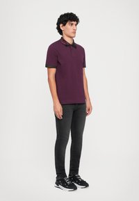 Homem de pé de lado, vestindo uma camisola polo roxa, jeans skinny pretos e tênis pretos com texto branco nos atacadores e solas.