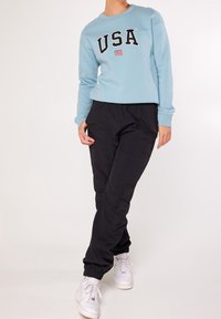 Lichtblauwe sweatshirt met de tekst "USA" en een vlaggraphic, gecombineerd met zwarte joggerbroek. Model draagt witte sneakers. Gladde stoftextuur.