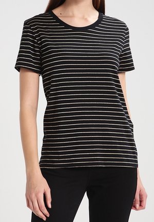 Vrouw draagt een zwart T-shirt met korte mouwen met dunne witte horizontale strepen, gecombineerd met een zwarte broek, staand tegen een effen achtergrond.