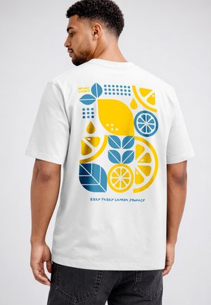 Mann mit weißem T-Shirt, darauf auffällige gelbe und blaue Zitrone, Blätter und geometrische Formen, mit dem Text "Easy Peasy Lemon Squeezy" auf dem Rücken.