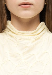 Κοντινό πλάνο στο κάτω μέρος του προσώπου και τον λαιμό ενός ατόμου που φοράει υφασμένο, κρεμ χρώματος μπλουζάκι με ψηλό λαιμό, με μακριά καστανά μαλλιά και μικρό χρυσό σκουλαρίκι.
