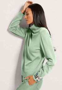 Frau in einem hellgrünen Rollkragenpullover und passenden Hosen steht im Profil mit einer Hand in der Tasche und der anderen Hand, die auf der Stirn ruht.