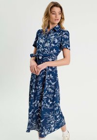 Kleid mit Blumenmuster in Marineblau, aus glatter Stoff gefertigt. Es hat einen kragenlosen Ausschnitt, kurze Ärmel, eine Knopfleiste und einen gebundenen Taillendetail.
