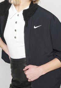 Svart zip-up jacka med strukturerad svart pälskrage, med en vit Nike-logotyp på bröstet. Bärs över en vit topp och svarta läderbyxor.