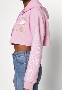 Osoba má na sobě růžovou krátkou mikinu s kapucí s grafikou Hello Kitty a nápisem na hrudi a rukávu, doplněnou modrými džínami.