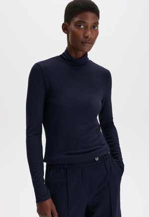 Femme aux cheveux courts portant un col roulé à manches longues bleu marine foncé et un pantalon assorti, debout devant un fond clair uni.