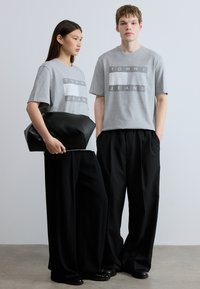 Γκρι κοντομάνικα μπλουζάκια με το λογότυπο «TOMMY JEANS», σε συνδυασμό με μαύρο παντελόνι φαρδιάς γραμμής και μαύρη τσάντα pouch. Απλός σχεδιασμός, υλικό από μείγμα βαμβακιού.