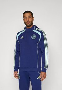 Tengerészkék kapucnis pulóver világoskék díszítésekkel és három csíkkal az ujjakon, elöl kerek logóval és húzózsinóros kapucnival.