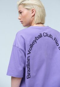 Lila Baumwoll-T-Shirt mit kurzen Ärmeln, auf dem schwarzer Text steht, der sich über den Rücken zieht und "Brazilian Volleyball Club, Rio de Janeiro" lautet.