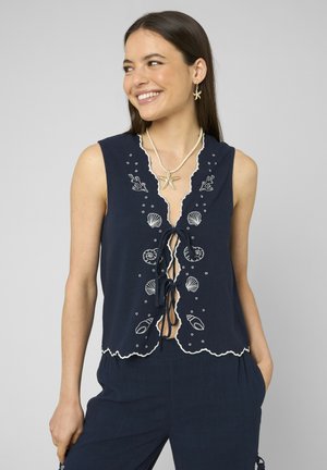Femme souriante portant un haut sans manches bleu marine avec une broderie blanche sur le thème de la mer, un collier étoile de mer, des boucles d’oreilles étoile de mer assorties, et un pantalon bleu marine.