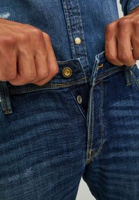 Jeans de denim azul con un corte relajado, que cuentan con cierres de botones dorados y plateados, costuras amarillas y un ligero patrón de desgaste a lo largo de los muslos.