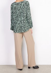Blusa verde con un motivo a foglia nera, maniche lunghe e scollatura rotonda, abbinata a pantaloni beige a gamba larga e scarpe nere.