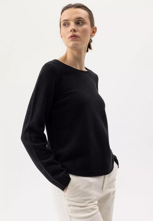 Giovane donna con maglione scuro e pantaloni chiari, mani in tasca, che guarda di lato su uno sfondo semplice.