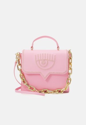 Mała różowa torebka na górny uchwyt, ze złotym łańcuszkiem na ramię i ozdobą w kształcie ćwieków w formie oka na przednim klapie, na białym tle.