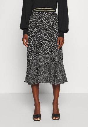 Kobieta nosząca czarno-białą, kwiatową spódnicę midi z falbaną na dole i złoto-paskowanym paskiem w talii, w połączeniu z czarnymi sandałami na obcasie z odkrytymi palcami.