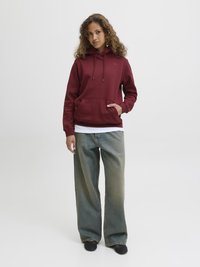 Bordeaux hettegenser med snorlås og frontlomme, sammen med vide, falmede blå jeans. Modellen har krøllete hår og står mot en nøytral bakgrunn.