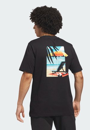 Hombre con cabello rizado que lleva una camiseta negra con una escena colorida de playa que muestra una tumbona y una sombrilla en la parte de atrás.