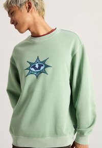 Felpa verde chiaro con collo a giro, caratterizzata da un design grafico blu e nero sul petto; maniche lunghe e polsini a costine.