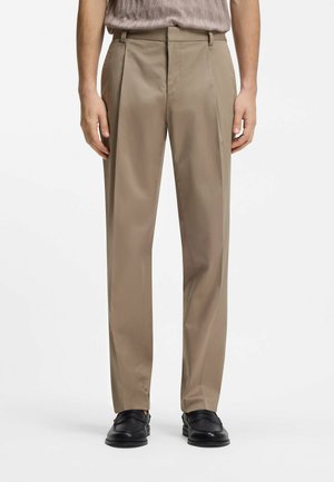 Beige bukser med lige ben, båret med sorte loafers og en lysebrun tekstureret skjorte stoppet ned i bukserne på en stående person.