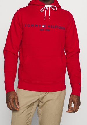 Muž v červené mikině Tommy Hilfiger s bílými šňůrkami a béžovými kalhotami stojí s rukama volně po stranách.
