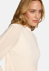 Femme blonde à la peau claire portant un pull crème avec des manches en maille ajourée, regardant légèrement de côté sur un fond blanc uni.