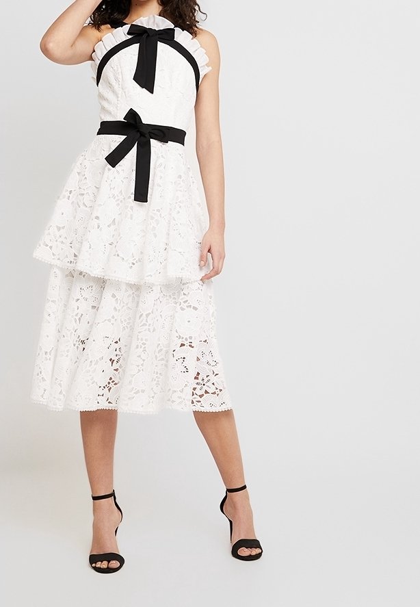 Femme portant une robe blanche en dentelle sans manches avec des détails de nœuds noirs au niveau du cou et de la taille, associée à des sandales à talons hauts noires.