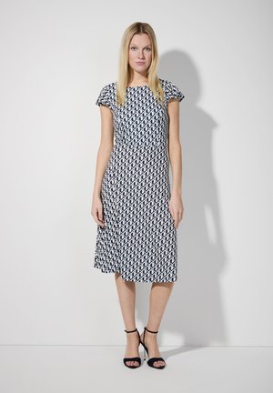 Navyblaues und weißes gemustertes Kleid mit kurzen Ärmeln, tailliertem Schnitt und weit schwingendem Rock. Getragen mit schwarzen, geschnallten High Heels.